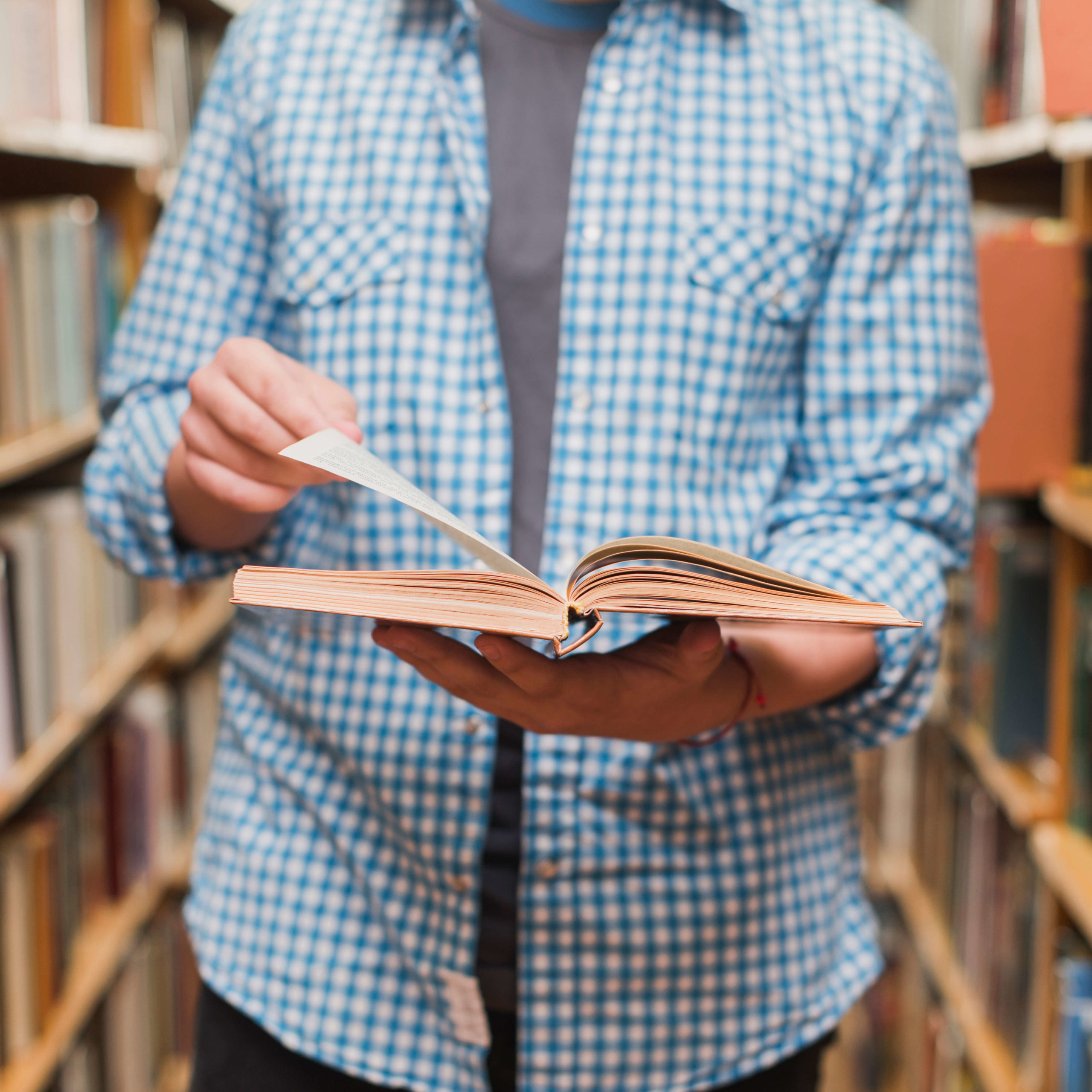 Hombre hojeando un libro de pie a espaldas de una biblioteca.